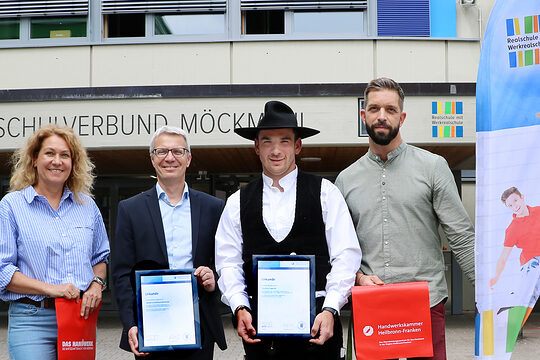 Eine Frau und drei Männer stehen vor einem Schulgebäude. Zwei der Männer halten eine Urkunde in den Händen.