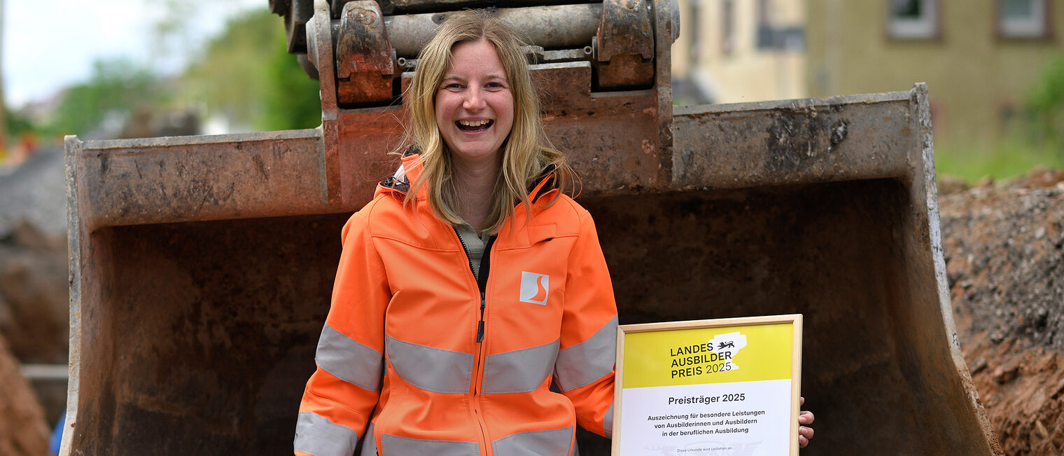 Eine Frau sitzt in einer Baggerschaufel. In der Hand hält sie eine Urkunde.
