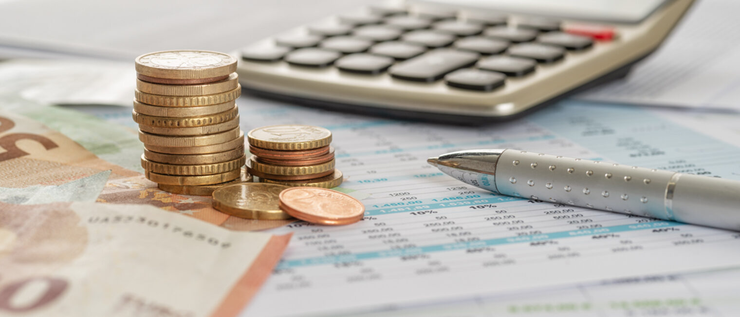 Handwerker erhalten Unterstützung in Sachen Gründungsfinanzierung. Auf einem Tisch liegen eine Tabelle, Geldscheine, Münzen, ein Taschenrechner und ein Kugelschreiber.