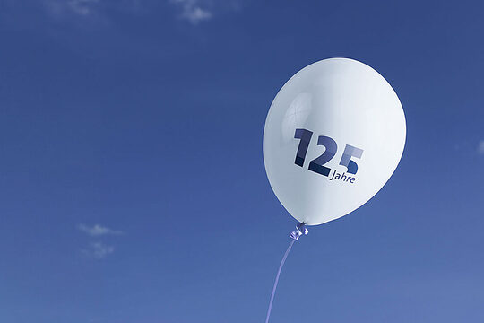 Ein weißer Luftballon mit der blauen Aufschrift "125 Jahre" schwebt rechts im Bild. Im Hintergrund ist der Himmel.