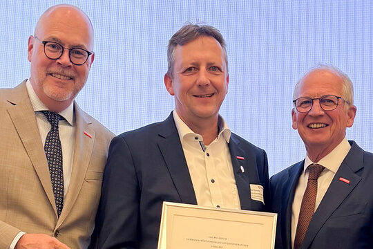 Drei Männer stehen nebeneinander. Der Mann in der Mitte hält eine Urkunde in den Händen.