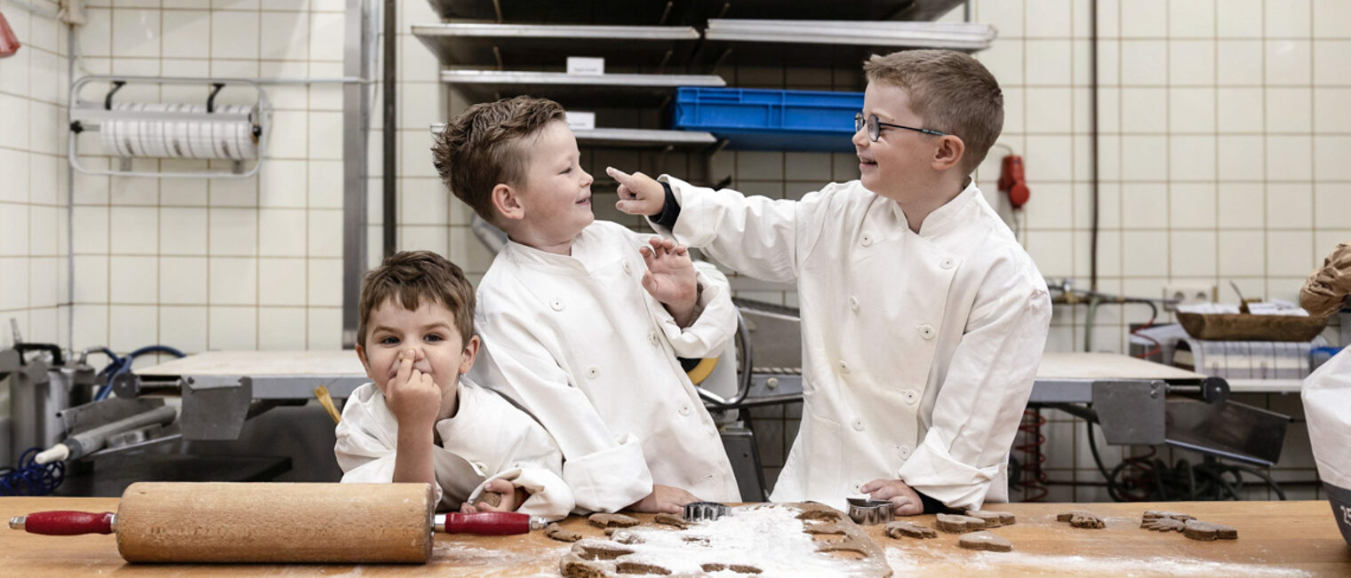 Der Kita-Wettbewerb des Handwerks verspricht allen Beteiligten viel Spaß. Drei Kinder stehen in einer Bäckerei hinter einem Tisch. Auf dem Tisch liegen ein Nudelholz und Teig.