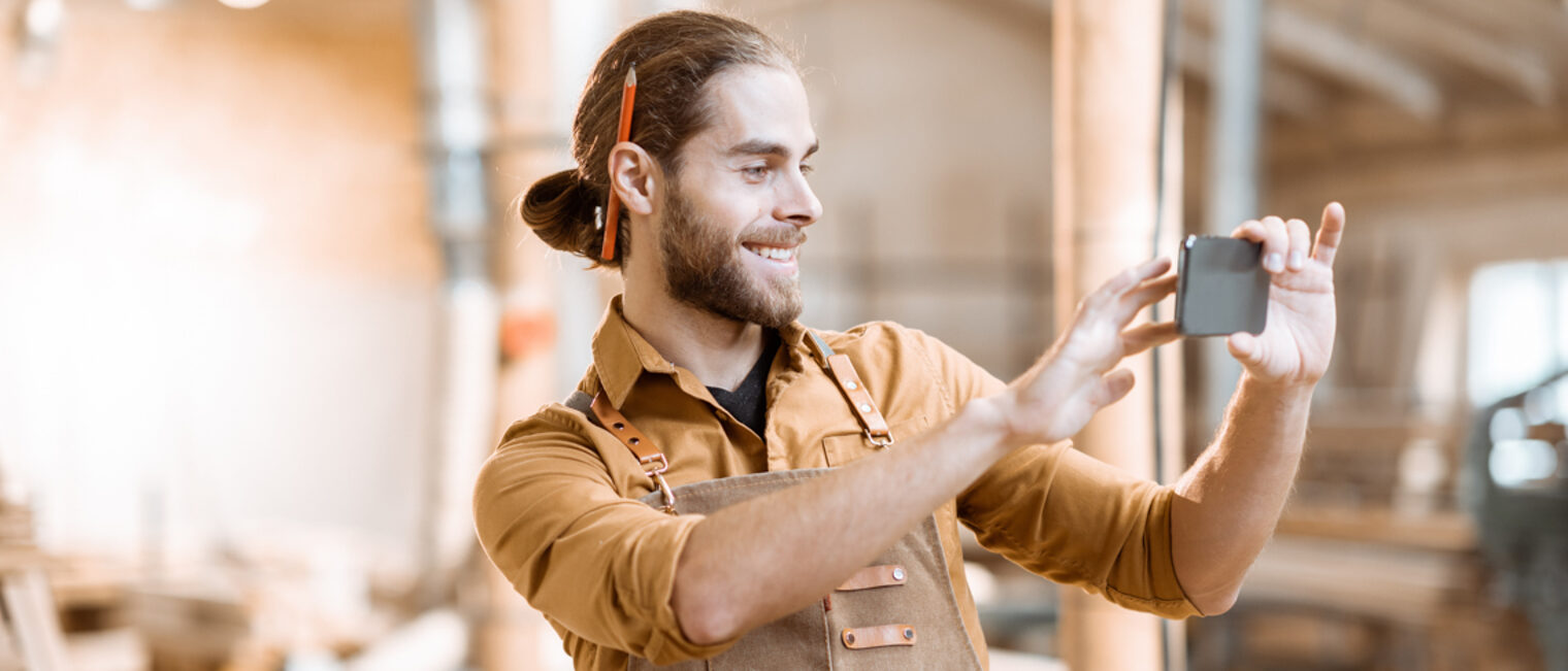 Ein Handwerker steht lächelnd in einer Werkstatt und filmt etwas mit seinem Smartphone.
