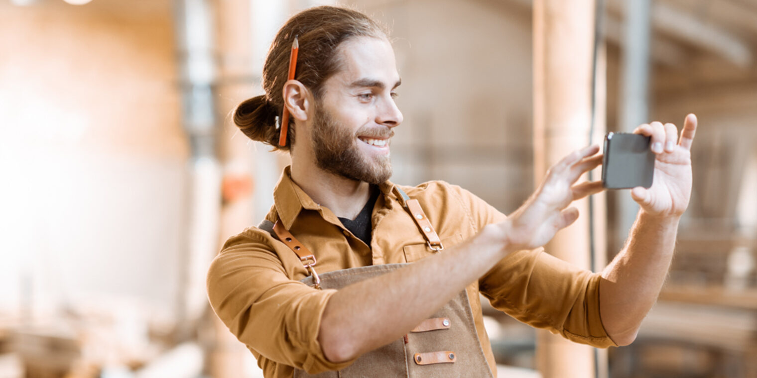 Ein Handwerker steht lächelnd in einer Werkstatt und filmt etwas mit seinem Smartphone.