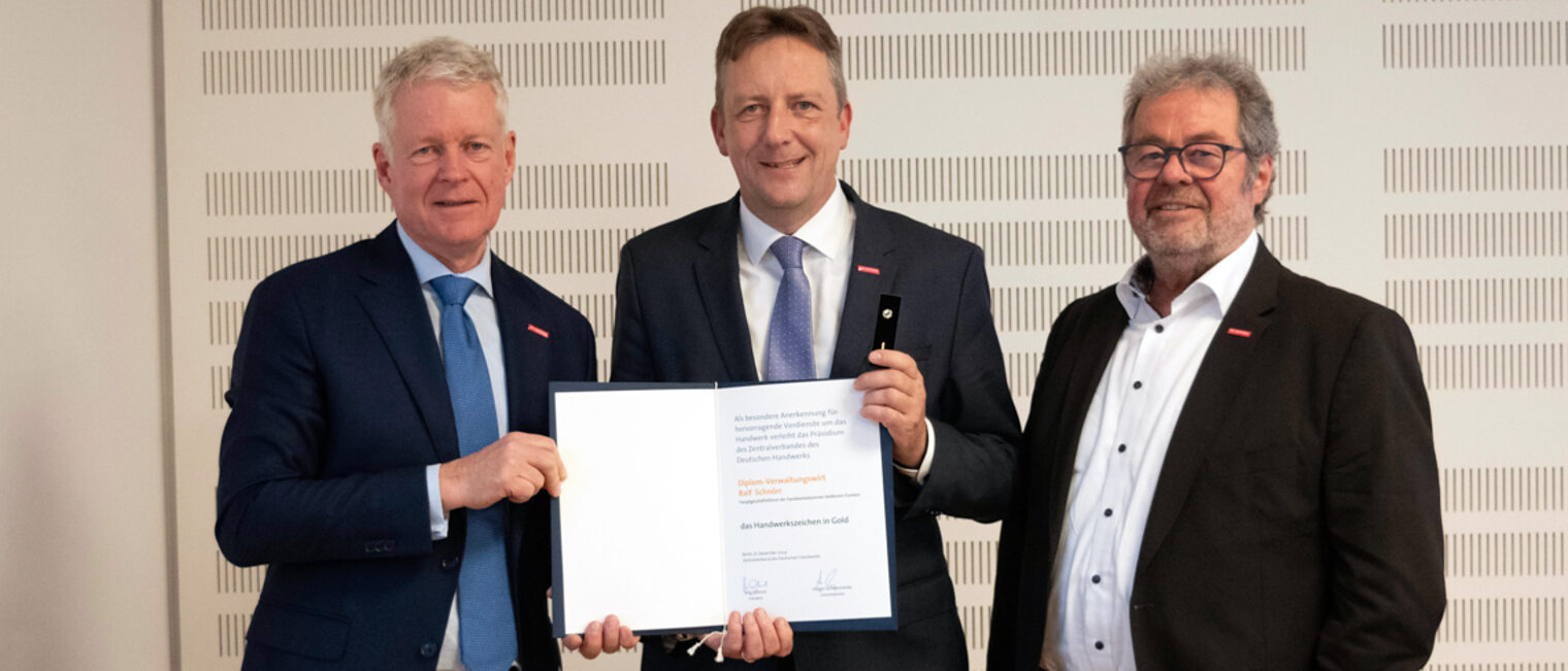 Drei Männer im Anzug, Mann in der Mitte und erhält eine Ehrungsurkunde.