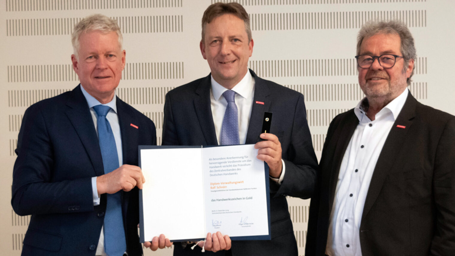 Drei Männer im Anzug, Mann in der Mitte und erhält eine Ehrungsurkunde.