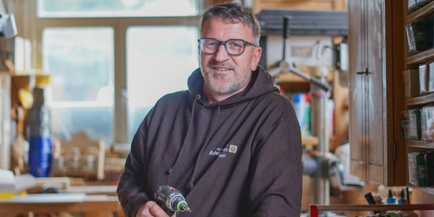 Ein Mann steht in einer Schreinerwerkstatt und hält einen Akku-Schreiber in der Hand. 