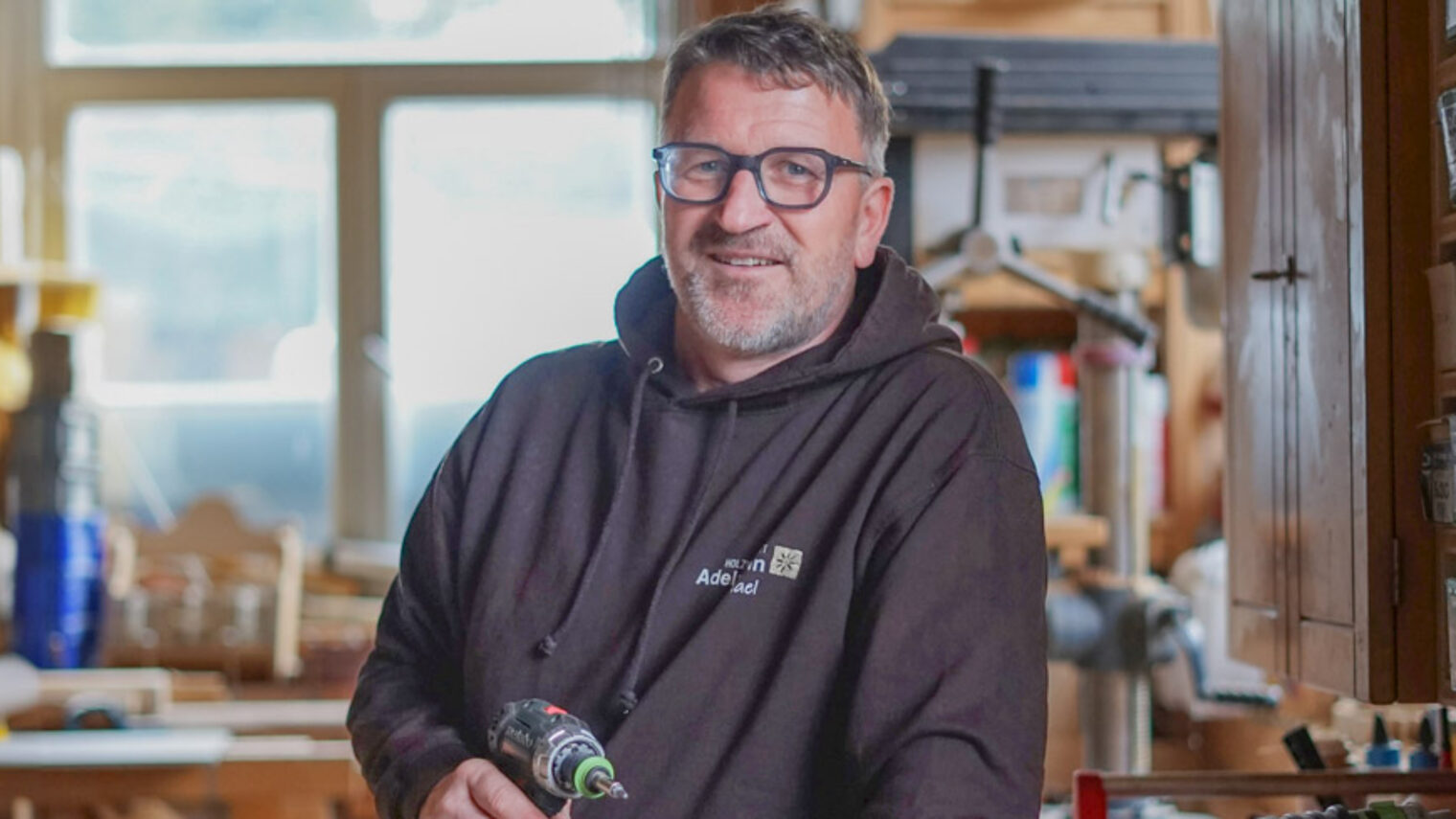 Ein Mann steht in einer Schreinerwerkstatt und hält einen Akku-Schreiber in der Hand. 