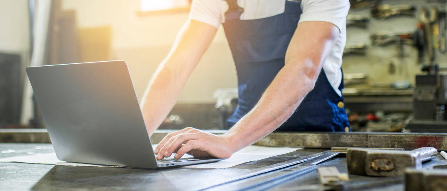 Ein Handwerker in Arbeitskleidung arbeitet an einem Laptop, der vor ihm auf dem Tisch steht.