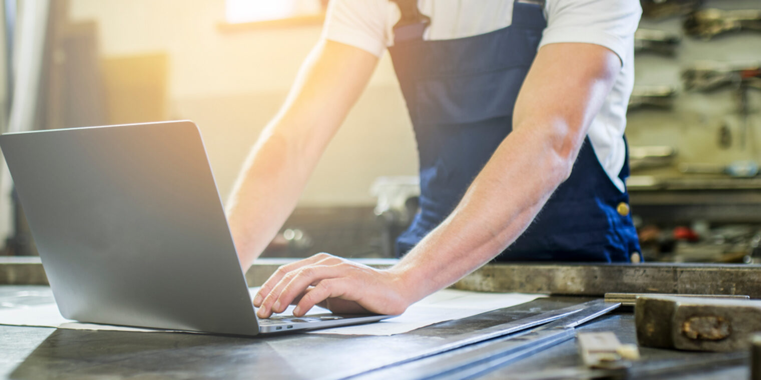 Ein Handwerker in Arbeitskleidung arbeitet an einem Laptop, der vor ihm auf dem Tisch steht.