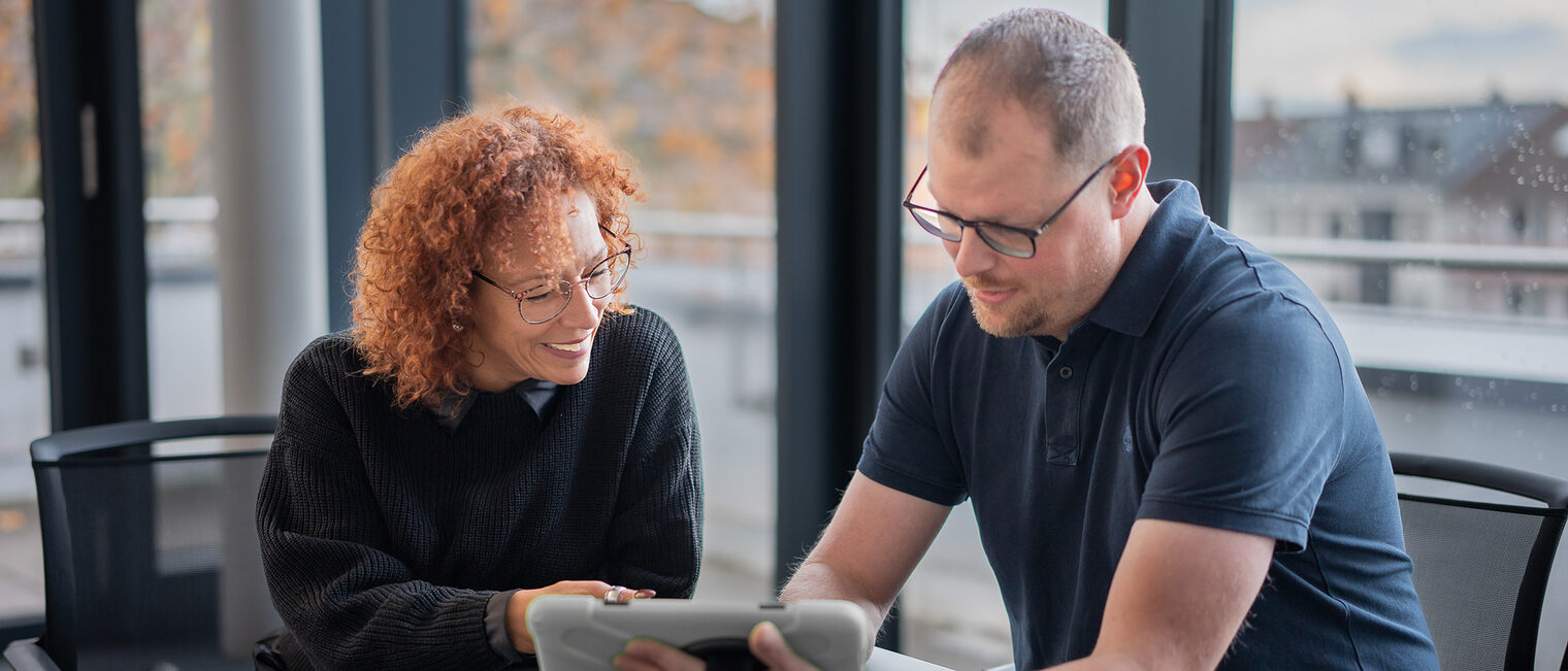 Eine Frau und ein Mann sitzen an einem Tisch und schauen gemeinsam auf ein Tablet.
