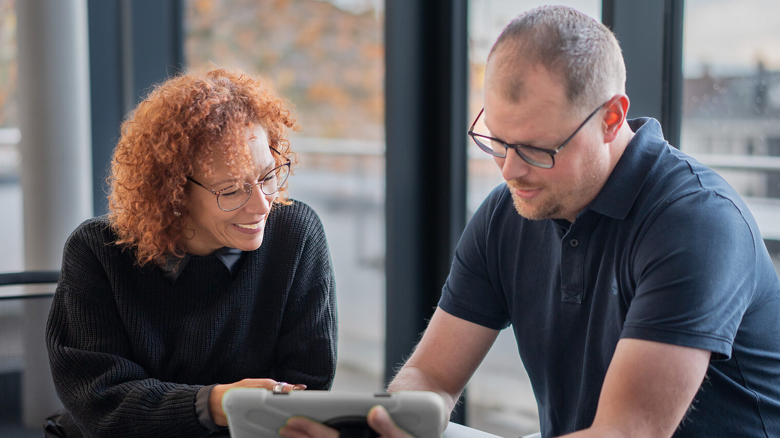 Eine Frau und ein Mann sitzen an einem Tisch und schauen gemeinsam auf ein Tablet.