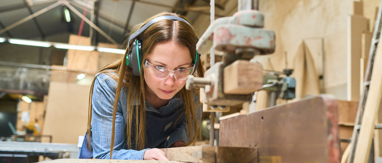 Eine Auszubildende zur Schreinerin steht an einer Werkbank und schneidet ein Stück Holz zurecht.