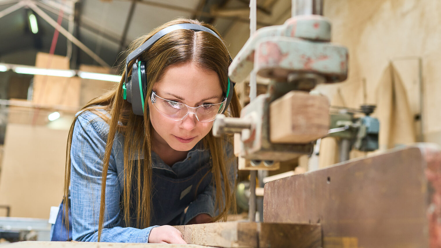 Eine Auszubildende zur Schreinerin steht an einer Werkbank und schneidet ein Stück Holz zurecht.