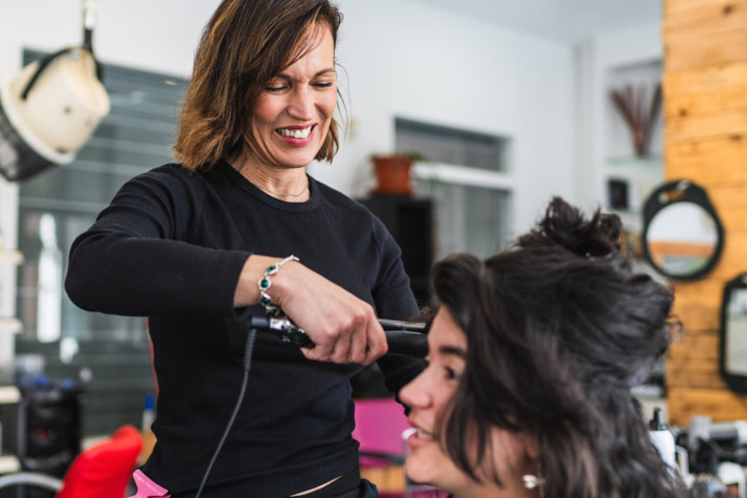 Eine Frau bearbeitet mit einem Glätteisen die Frisur einer Kundin einem Friseursalon. 