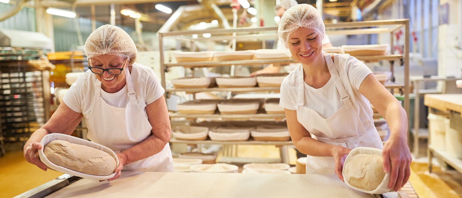 In einer Backstube stehen zwei Bäckerinnen. Gemeinsam bereiten sie Brotlaibe zum Backen vor.