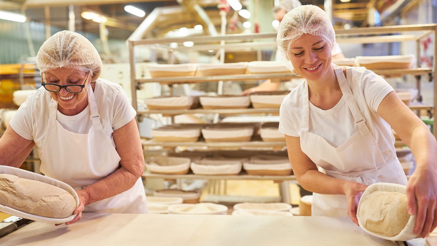 In einer Backstube stehen zwei Bäckerinnen. Gemeinsam bereiten sie Brotlaibe zum Backen vor.