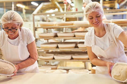 In einer Backstube stehen zwei Bäckerinnen. Gemeinsam bereiten sie Brotlaibe zum Backen vor.