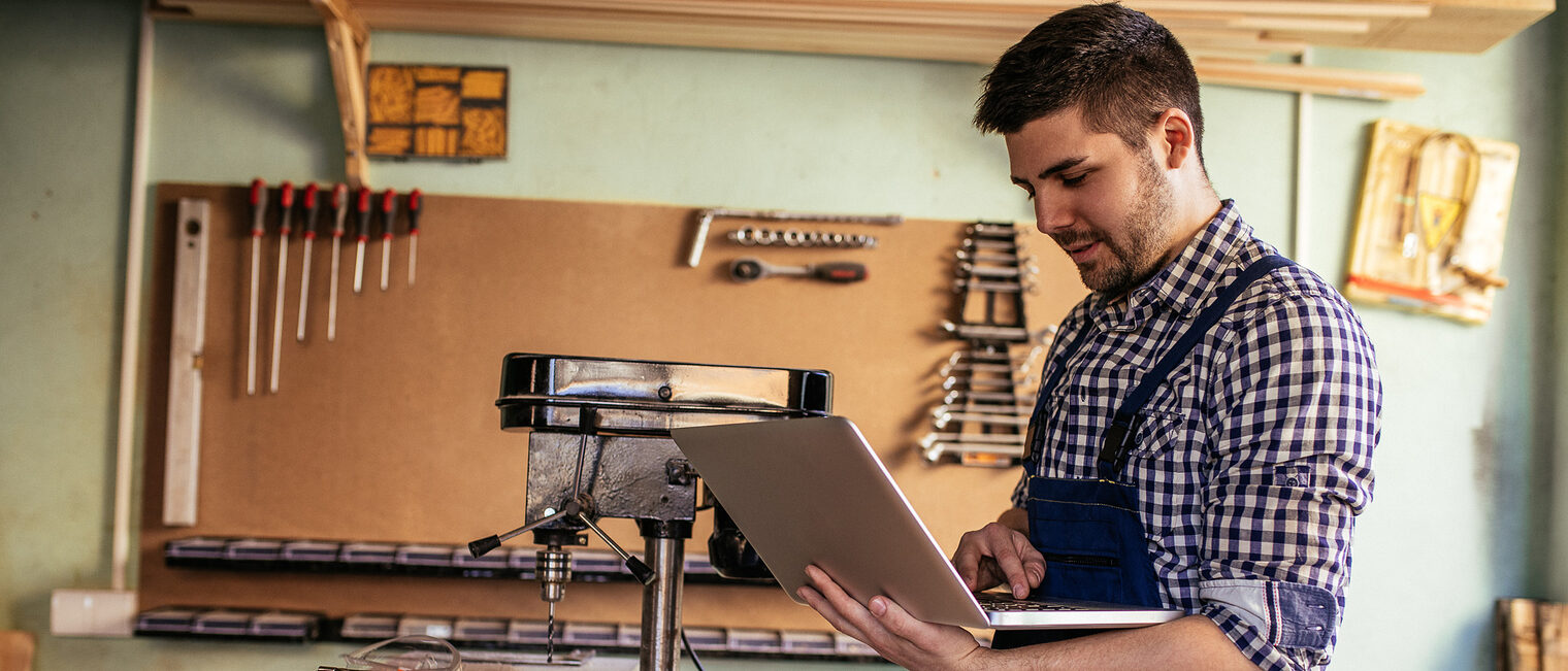 Ein junger Handwerker steht in einer Werkstatt. In seinen Händen hält er einen Laptop.