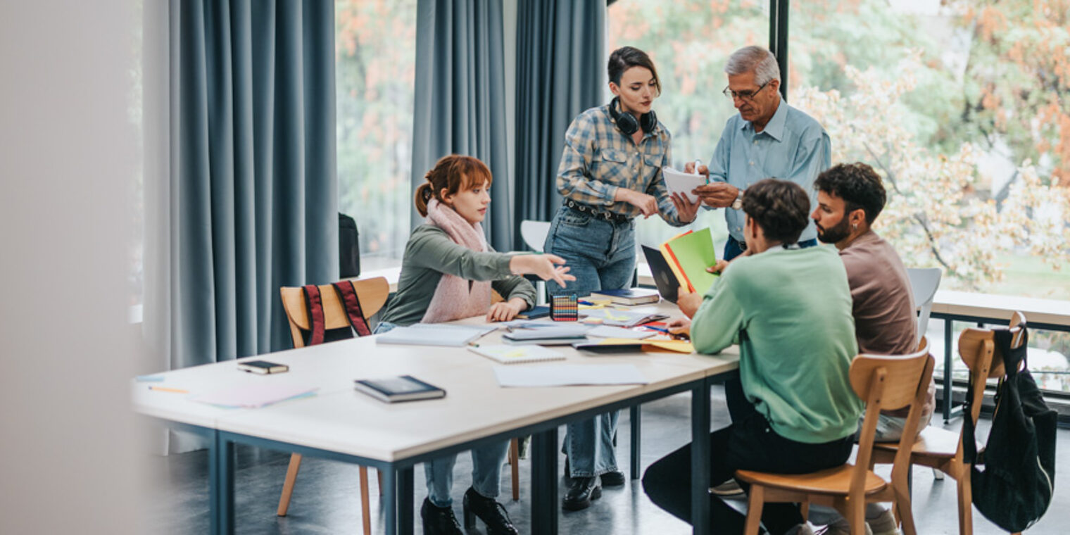 Ein Studententeam sitzt zur Teambesprechung am Tsch an der Hochschule und sprechen mit einem erfahrenem Mann.