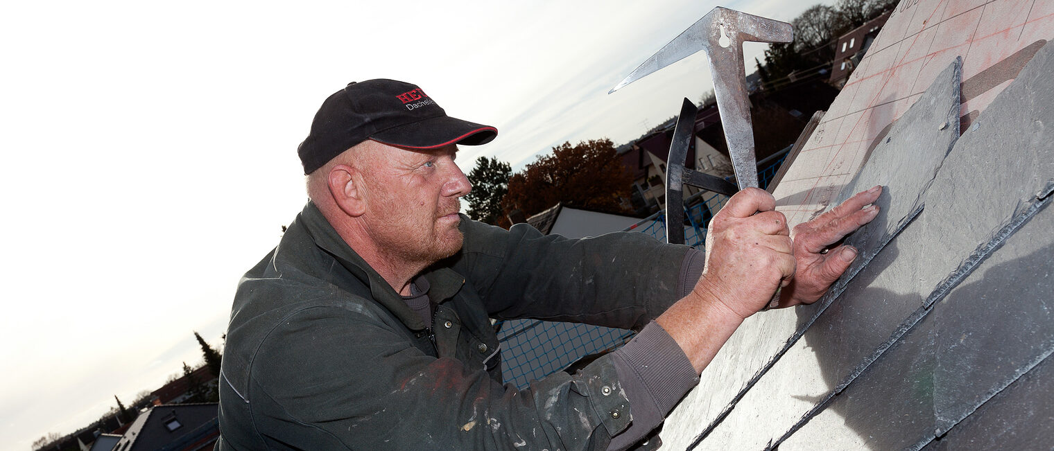 Ein Dachdecker hält einen Hammer in der Hand, mit dem er Schindeln auf einem Schrägdach befestigt.