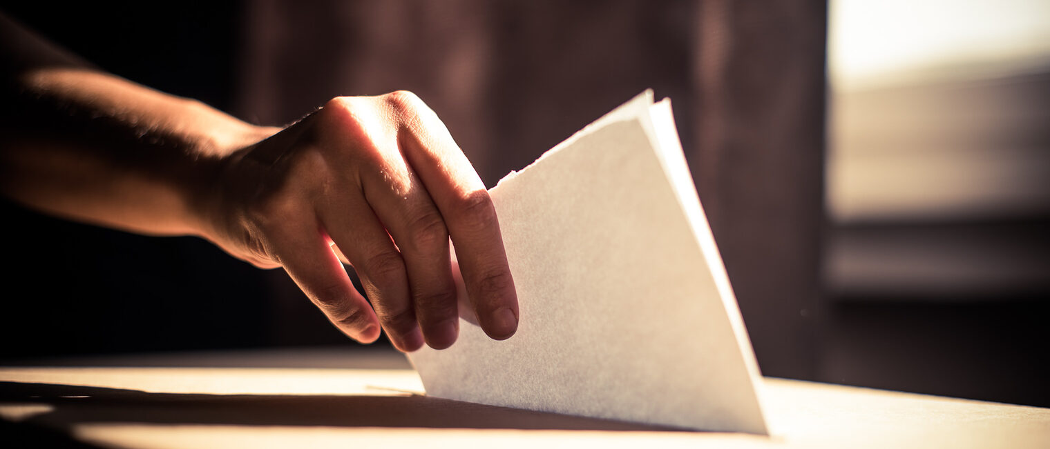 In Nahaufnahme ist zu sehen, wie jemand einen Stimmzettel in eine Wahlurne wirft.