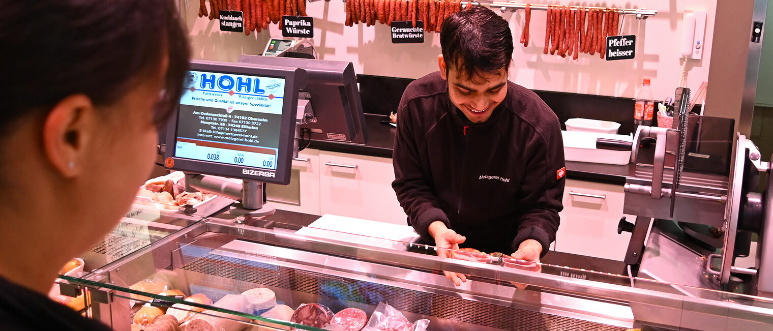 Ein Fleischereifachverkäufer-Azubi bedient in einer Metzgerei eine Kundin.