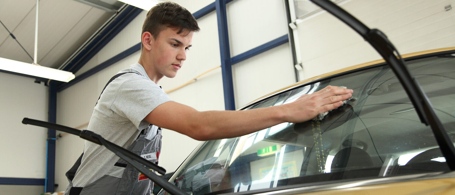 Ein junger Mann, der eine Ausbildung zum Fahrzeuglackierer absolviert, poliert die Scheibe eines Autos.