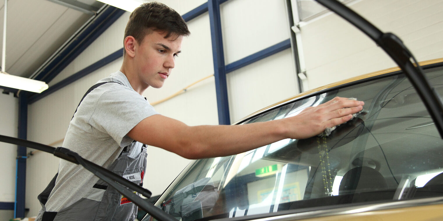 Ein junger Mann, der eine Ausbildung zum Fahrzeuglackierer absolviert, poliert die Scheibe eines Autos.