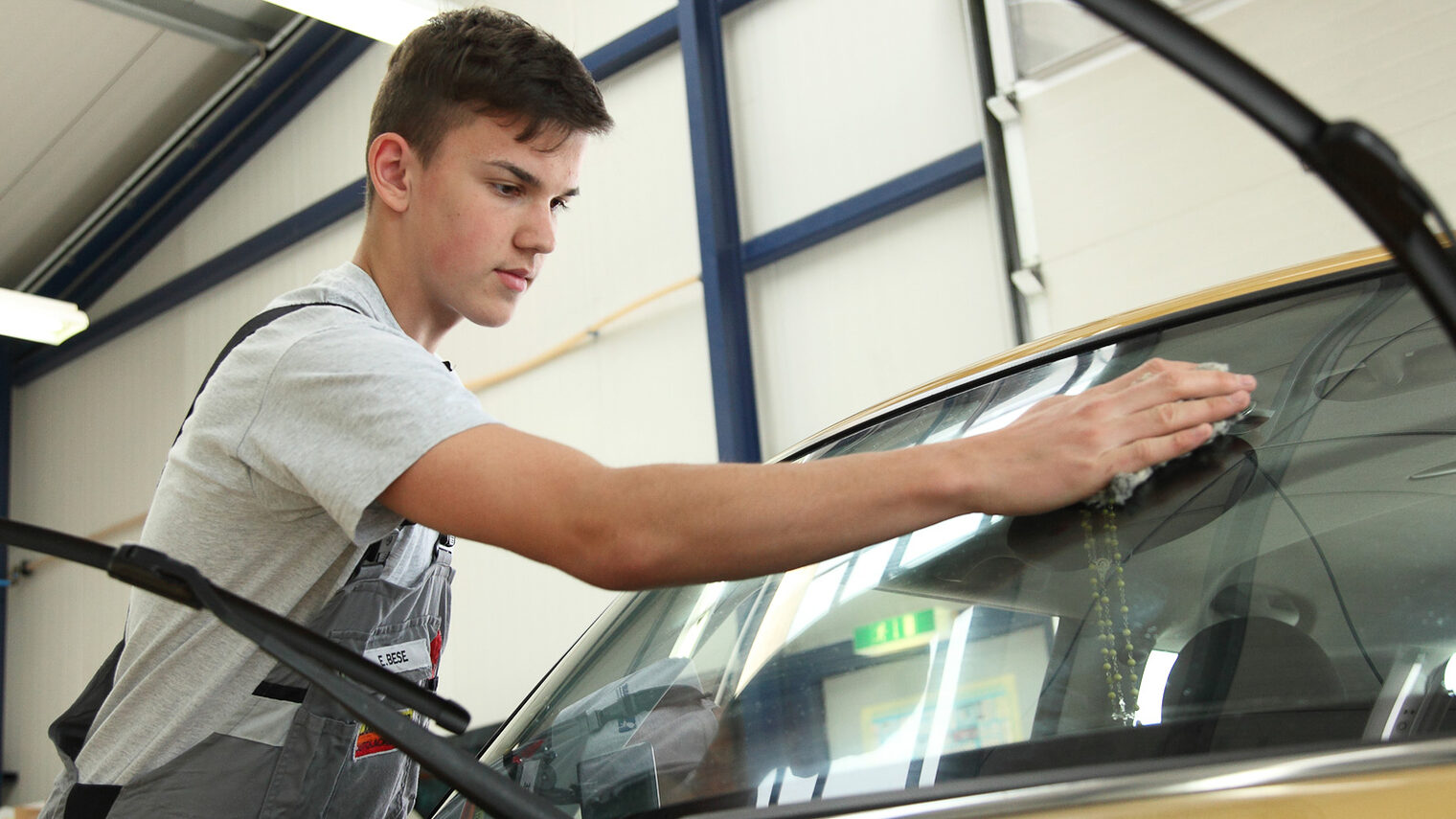 Ein junger Mann, der eine Ausbildung zum Fahrzeuglackierer absolviert, poliert die Scheibe eines Autos.