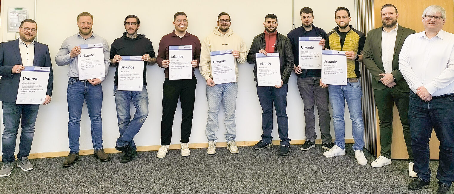 Zehn Männer stehen vor einer Wand und halten Urkunden in der Hand. 