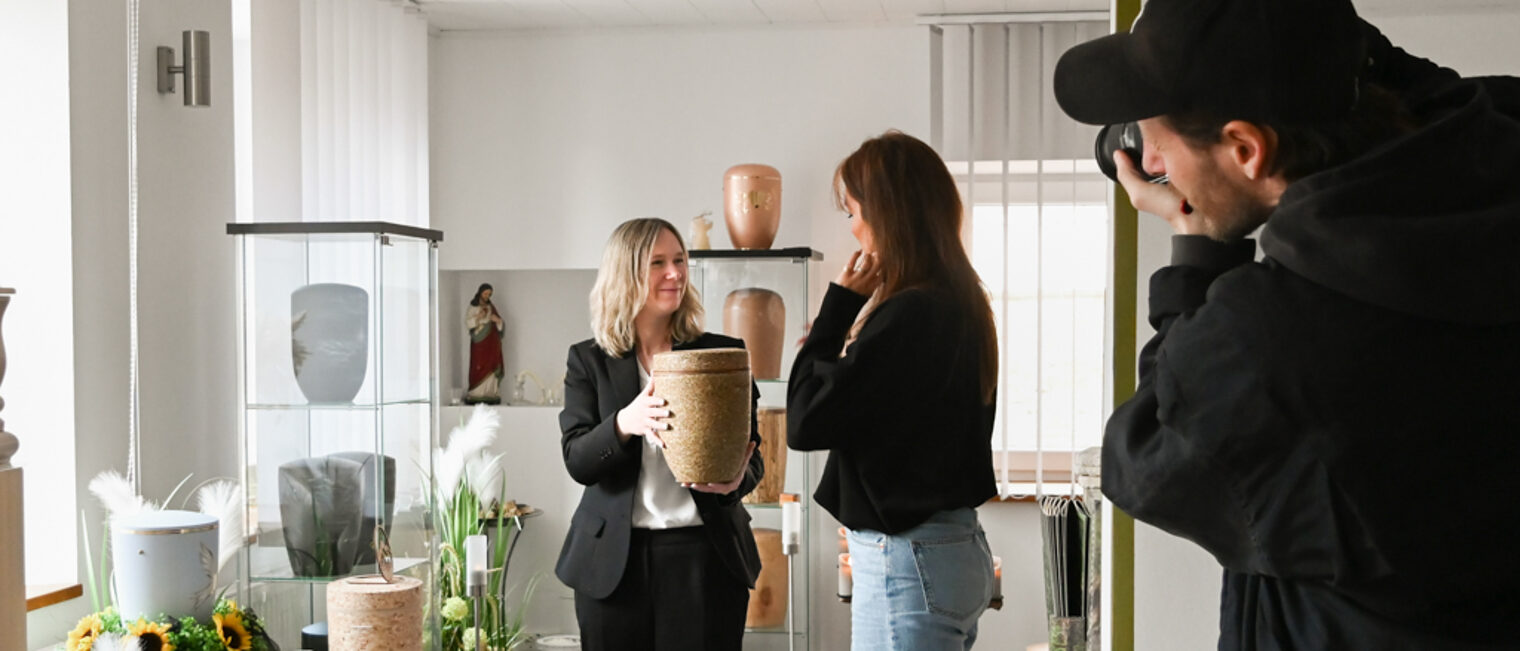 Bestatterin im Gespräch, sie zeigt eine Urne aus Kräutern für Naturbestattungen und wird dabei fotografiert