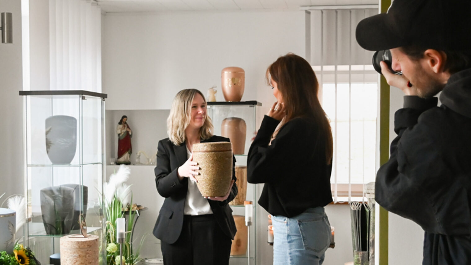 Bestatterin im Gespräch, sie zeigt eine Urne aus Kräutern für Naturbestattungen und wird dabei fotografiert