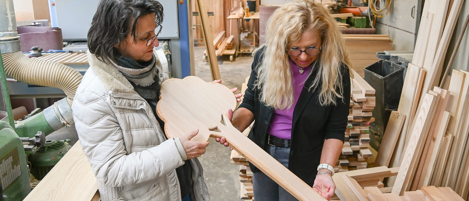 In einer Holzwerkstatt stehen zwei Frauen und schauen sich handgefertigten Grabschmuck aus Holz an.