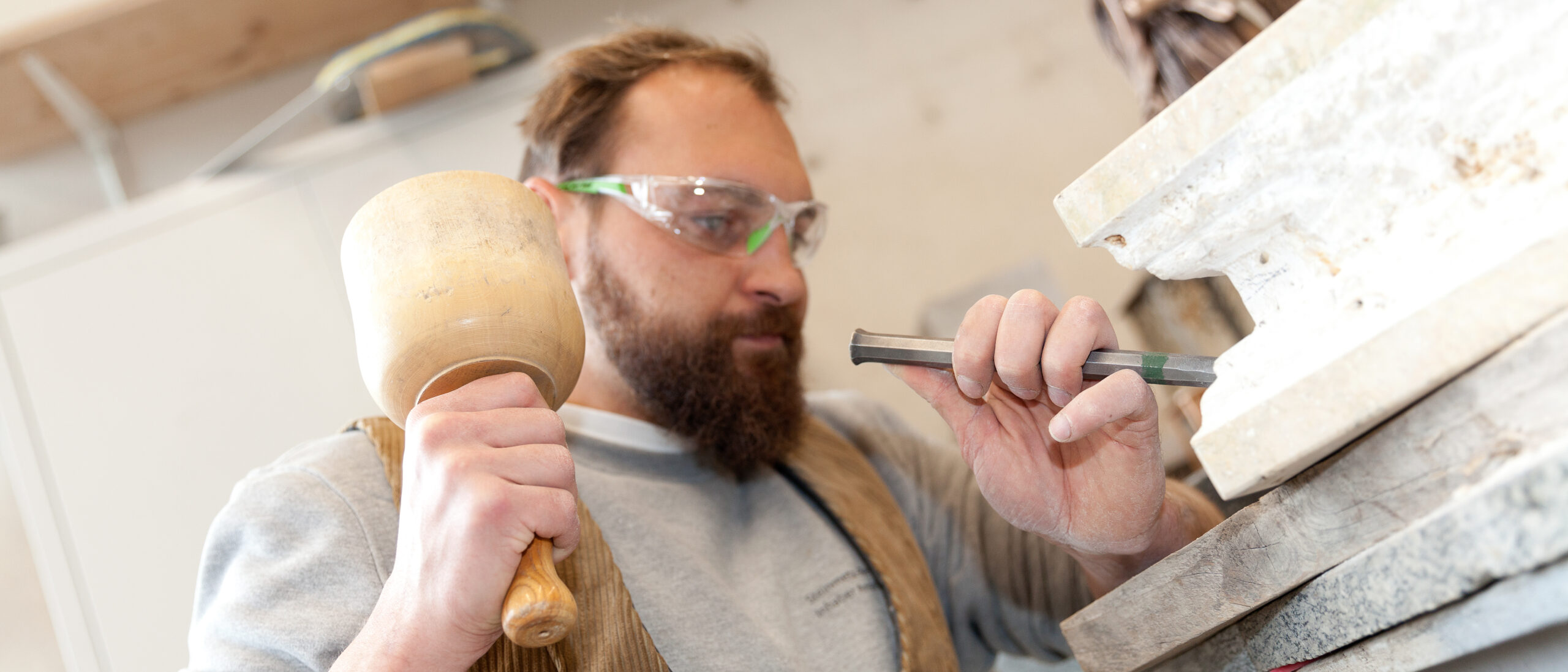 Ein Mann trägt eine Schutzbrille und bearbeitet mit Hammer und Meißel eine Steinplatte.