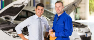Ein Mann mit Krawatte und ein Mann im Blaumann stehen vor zwei Autos, deren Motorhauben geöffnet sind.