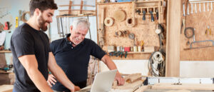 Junior und Senior Handwerker schauen in einer Schreinerei gemeinsam etwas am Laptop an und sind zufrieden.