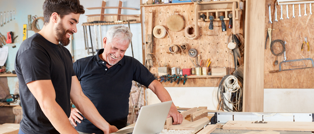 Junior und Senior Handwerker schauen in einer Schreinerei gemeinsam etwas am Laptop an und sind zufrieden.