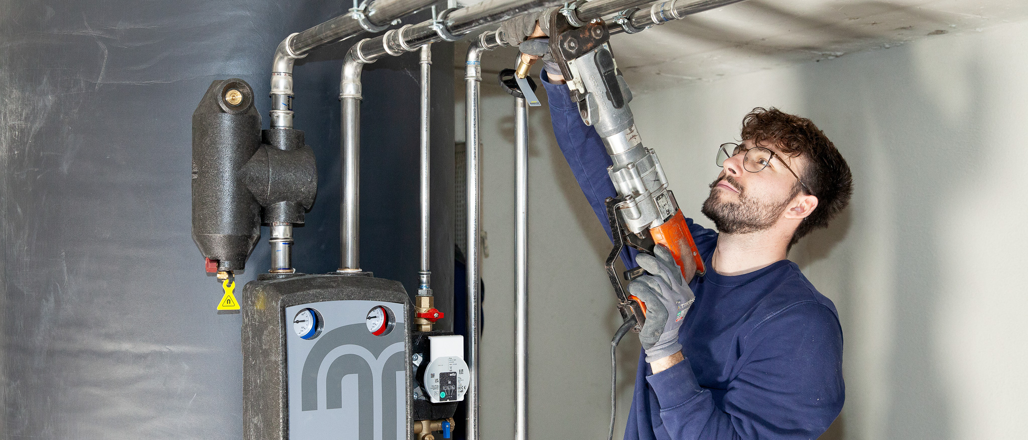 Ein Anlagenmechaniker für Sanitär-, Heizungs- und Klimatechnik arbeitet mit seinem Werkzeug an den Rohren einer Heizungsanlage.