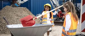 Zwei Kleine Mädchen mit Warnweste stehen auf einer Baustellen und schaufeln Sand in eine Schubkarre.