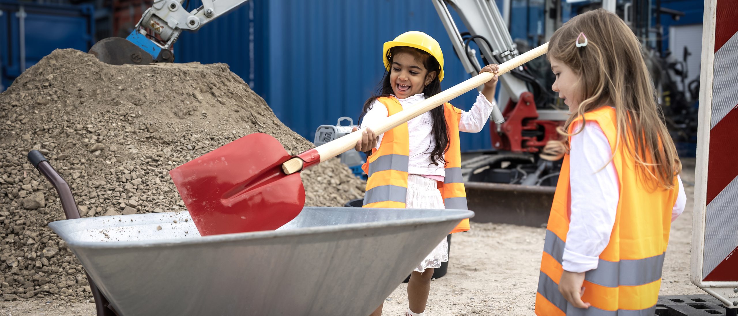 Zwei Kleine Mädchen mit Warnweste stehen auf einer Baustellen und schaufeln Sand in eine Schubkarre.